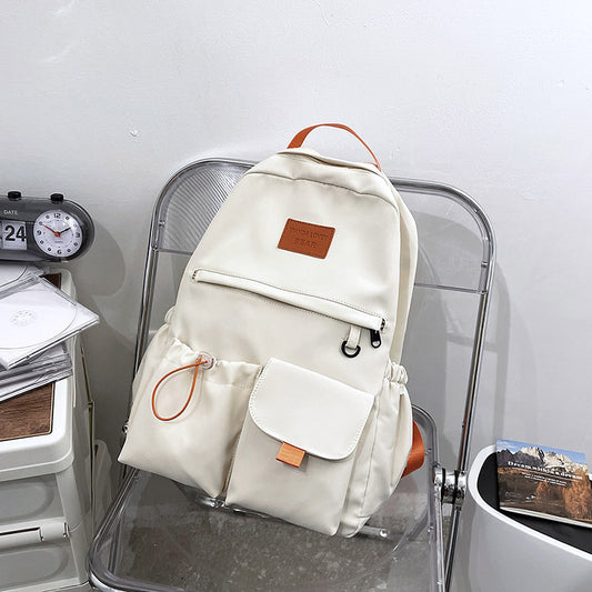 Cream canvas backpack with orange accents, front pockets, and leather patch, styled on clear chair for school or travel