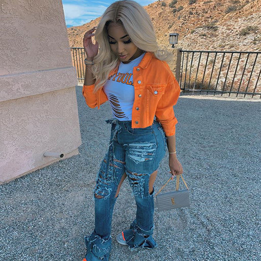 Woman in orange denim jacket, ripped jeans, and white graphic tee, holding gray handbag outdoors