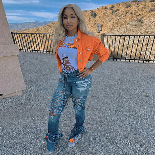 Woman in orange denim jacket, ripped jeans, and distressed tee, posing outdoors with mountain view