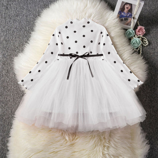 White polka dot girls dress with tulle skirt and black ribbon belt, styled on fluffy rug with photo and flowers