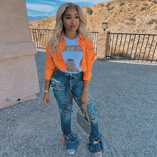 Woman in orange denim jacket, ripped jeans, and distressed top, holding gray handbag outdoors
