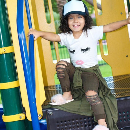 Child in playful outfit with eyelash print tee, ripped jeans, and cap at playground