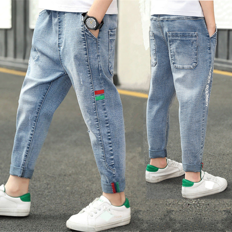 Kids' light blue denim joggers with red-green stripe, cuffed hems, and side lettering, paired with white sneakers