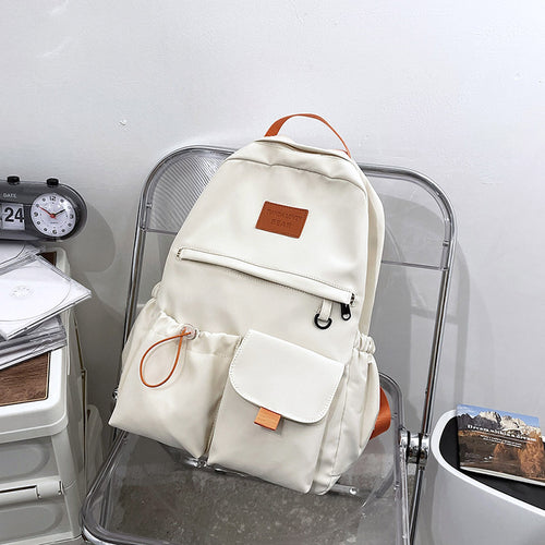 Cream canvas backpack with orange accents, front pockets, and leather patch, styled on clear chair for school or travel
