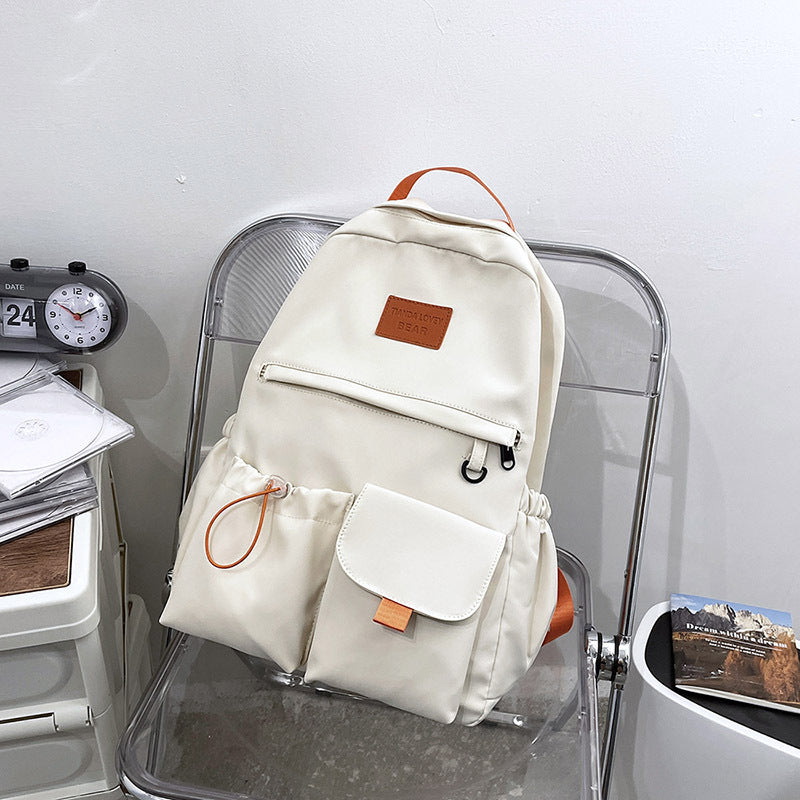 Cream canvas backpack with orange accents, front pockets, and leather patch, styled on clear chair for school or travel