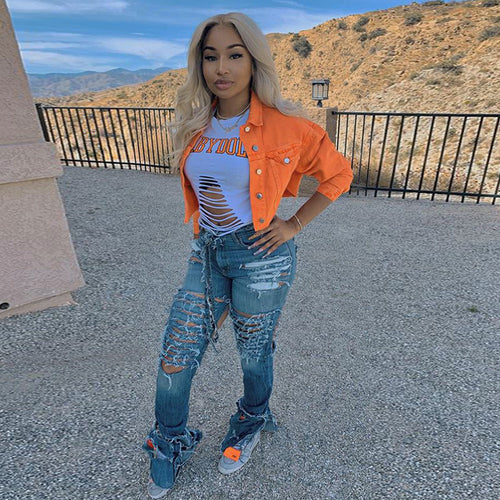 Woman in orange denim jacket, ripped jeans, and distressed tee, posing outdoors with mountain view
