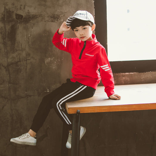 Boy in red zip-up jacket, black stripe pants, white sneakers, and cap, sitting on table
