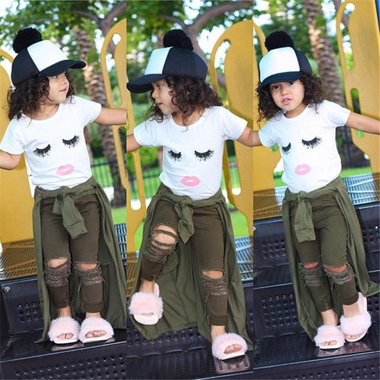 Toddler girl in cute eyelash print tee, ripped olive pants, fluffy slippers & pom-pom hat, playful outdoor pose