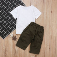 White kids t-shirt with olive green shorts, laid on wood floor beside checkered mat and star decor