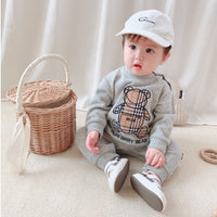 Baby in gray Burberry Bear sweatshirt, white cap, and sneakers, sitting beside wicker basket