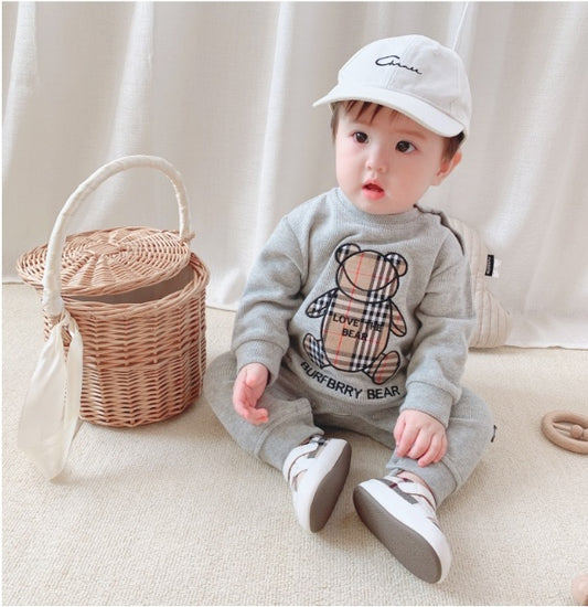 Baby in gray Burberry Bear sweatshirt, white cap, and sneakers, sitting beside wicker basket