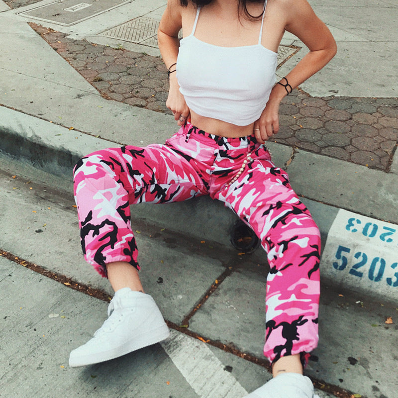Woman in pink camo pants, white crop top, and sneakers posing on urban steps