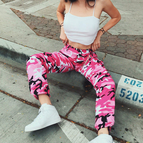 Woman in pink camo pants, white crop top, and sneakers posing on urban steps