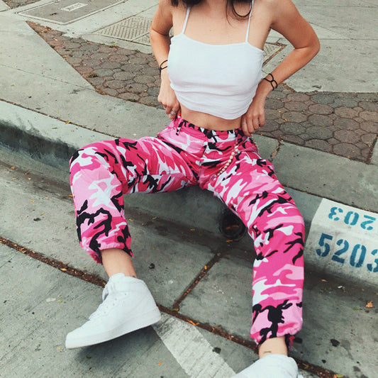 Woman in pink camo pants, white crop top, and sneakers posing on urban steps
