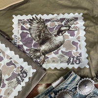 Olive green Duck Stamp graphic tee by Wild Junkie featuring a detailed duck stamp design on a casual men's graphic T-shirt.